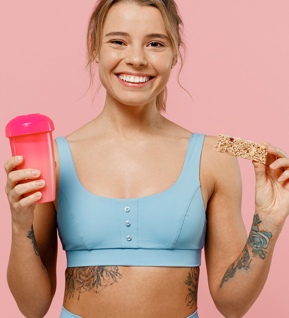 Femme sportive souriante tenant un shaker et une barre protéinée, symbolisant une alimentation équilibrée pour le sport