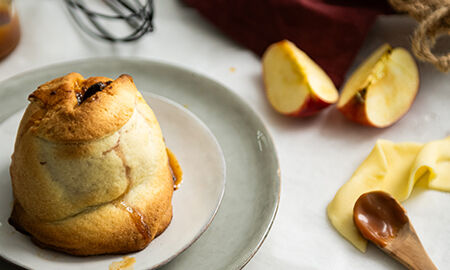Douillon de pomme doré avec caramel, accompagné de quartiers de pomme et d’une cuillère en bois sur une table lumineuse